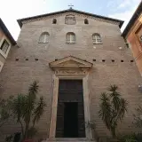 Facciata in mattoni di Santa Prassede a Roma, con un grande portale in legno e piante verdi.