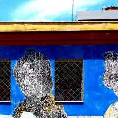 Volti stilizzati in bianco e nero su un muro blu acceso, parte di un **murales ostiense**. Le finestre con grate interrompono la composizione.