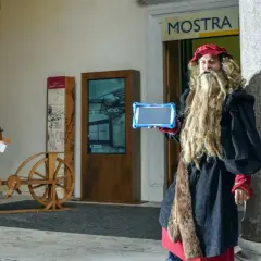 Uomo vestito da Leonardo da Vinci con tablet, pronto per una caccia al tesoro a Roma, vicino a un'antica bicicletta di legno.