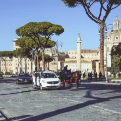 Carrozza trainata da cavalli, taxi e autobus sfilano su un lastricato romano, con la cupola di una chiesa e un monumento antico sullo sfondo, nella zona Battistini Roma.