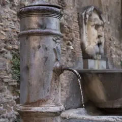 Fontanella romana con un nasone che eroga acqua fresca. Sullo sfondo, un antico volto scolpito.