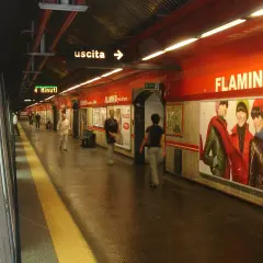 Stazione della metro Roma Nord, banchina con persone in attesa, cartelloni pubblicitari e indicazioni per l'uscita.