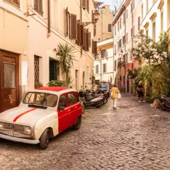 Un'auto d'epoca bianca e rossa parcheggiata in un vicolo acciottolato del **quartier Trastevere**. Persone passeggiano, un cane annusa l'aria.