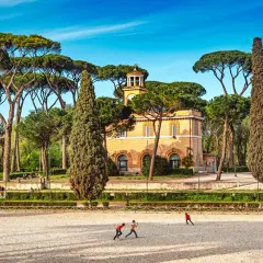 Un edificio giallo con una torre dell'orologio, circondato da pini marittimi e cipressi. Bambini giocano in un'area sabbiosa. Ecco cosa vedere ai Parioli.