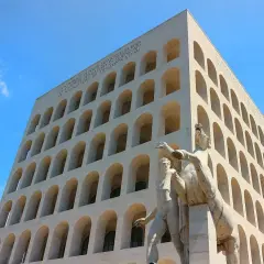 Palazzo Mussolini, con la sua facciata ad archi sovrapposti, domina il cielo azzurro. Una scultura di uomini e cavalli si erge in primo piano.
