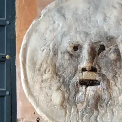 La Bocca della Verità, un volto scolpito nella pietra, sembra pronto a giudicare chiunque osi mentire. Accanto, una porta scura.
