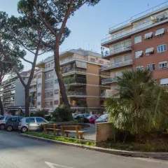 Edifici residenziali e pini marittimi nel quartiere Balduina a Roma. Auto parcheggiate lungo la strada, panchine e vegetazione rigogliosa.