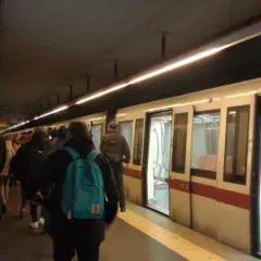 Persone in attesa sul binario della metro domenica, con un treno bianco e rosso fermo con le porte aperte.
