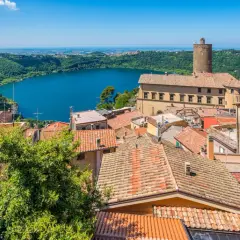 Panoramica dei colli di Roma oggi: un lago azzurro circondato da colline verdi e un borgo con tetti rossi e una torre medievale.
