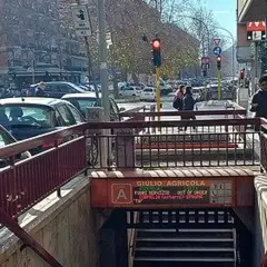 Ingresso della fermata Giulio Agricola della metro A, con persone che camminano e traffico in strada.