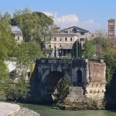 Antico **ponte rotto a Roma**, con rovine e vegetazione che si arrampicano sui resti. In lontananza, edifici storici e una torre campanaria.