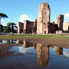 Le maestose rovine delle **Terme di Caracalla** si specchiano in una pozza d'acqua, sotto un cielo azzurro con nuvole bianche.
