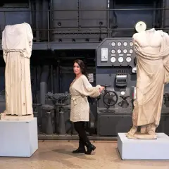 Donna in abito scintillante tra statue classiche e macchinari industriali, forse nel vecchio gasometro.