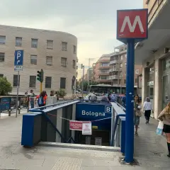 Ingresso della metro Bologna a Roma, con insegna rossa e bianca. Persone camminano sul marciapiede.
