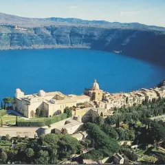 Un borgo pittoresco si affaccia su un lago blu intenso, incastonato tra i verdi **colli romani**. Un complesso di edifici storici domina la scena.
