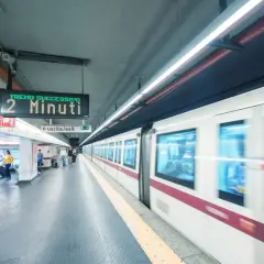 Stazione metro Roma: un treno in arrivo tra 2 minuti. Persone attendono sul binario, un cartello indica "uscita/exit".