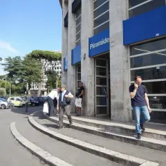 Uscita della metro Piramide a Roma Ostiense. Persone entrano ed escono dall'edificio, mentre auto sono parcheggiate nelle vicinanze.