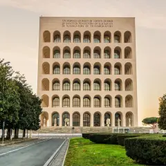 Edificio con arcate sovrapposte, simile a una stazione metro EUR Magliana, con iscrizione in latino e alberi verdi.