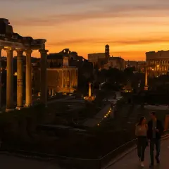 Coppia passeggia al tramonto tra le rovine del Foro Romano, con il Colosseo illuminato sullo sfondo. Un'atmosfera magica, ideale per scoprire Roma nel periodo migliore.