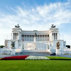Il Vittoriano, monumento imponente, si erge maestoso. Vicino alla fermata metro Altare della Patria, un prato verde e fiori rossi e bianchi creano un contrasto vibrante.