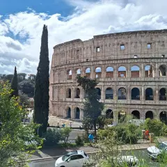Le maestose **colonne di Roma** del Colosseo si stagliano contro un cielo azzurro con nuvole bianche. Alberi verdi e un'auto bianca completano la scena.