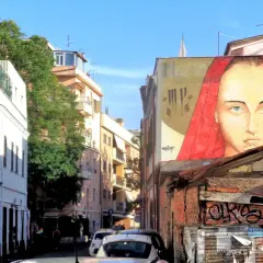 Un grande volto femminile con un velo rosso domina un edificio nel quartiere Pigneto. Accanto, un occhio gigante osserva la strada.