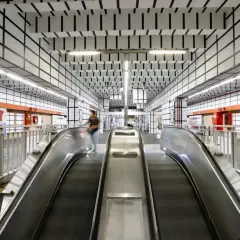 Scale mobili nella moderna stazione metro Vittorio Emanuele a Roma, con soffitto a griglia nera su bianco e opere d'arte colorate alle pareti.