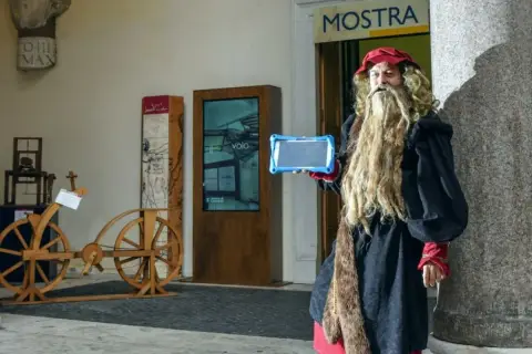 Uomo vestito da Leonardo da Vinci con tablet, pronto per una caccia al tesoro a Roma, vicino a un'antica bicicletta di legno.