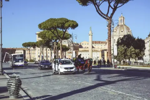 Carrozza trainata da cavalli, taxi e autobus sfilano su un lastricato romano, con la cupola di una chiesa e un monumento antico sullo sfondo, nella zona Battistini Roma.