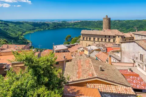 Panoramica dei colli di Roma oggi: un lago azzurro circondato da colline verdi e un borgo con tetti rossi e una torre medievale.