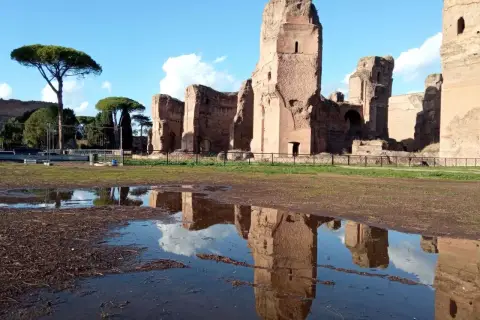 Le maestose rovine delle **Terme di Caracalla** si specchiano in una pozza d'acqua, sotto un cielo azzurro con nuvole bianche.