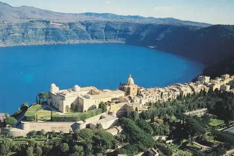 Un borgo pittoresco si affaccia su un lago blu intenso, incastonato tra i verdi **colli romani**. Un complesso di edifici storici domina la scena.