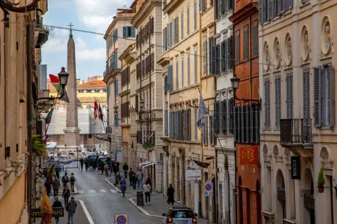 Una strada romana con palazzi storici, un obelisco e gente che passeggia. Si intuisce quanto è lunga Via del Corso, con la sua prospettiva che si perde all'orizzonte.