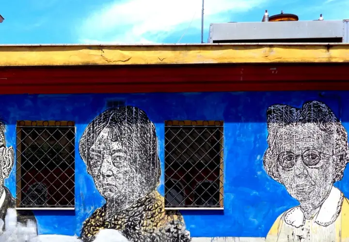 Volti stilizzati in bianco e nero su un muro blu acceso, parte di un **murales ostiense**. Le finestre con grate interrompono la composizione.