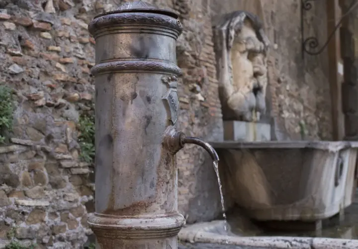 Fontanella romana con un nasone che eroga acqua fresca. Sullo sfondo, un antico volto scolpito.