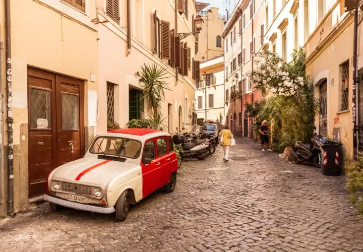 Un'auto d'epoca bianca e rossa parcheggiata in un vicolo acciottolato del **quartier Trastevere**. Persone passeggiano, un cane annusa l'aria.