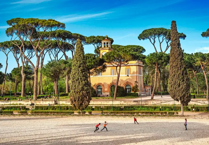 Un edificio giallo con una torre dell'orologio, circondato da pini marittimi e cipressi. Bambini giocano in un'area sabbiosa. Ecco cosa vedere ai Parioli.