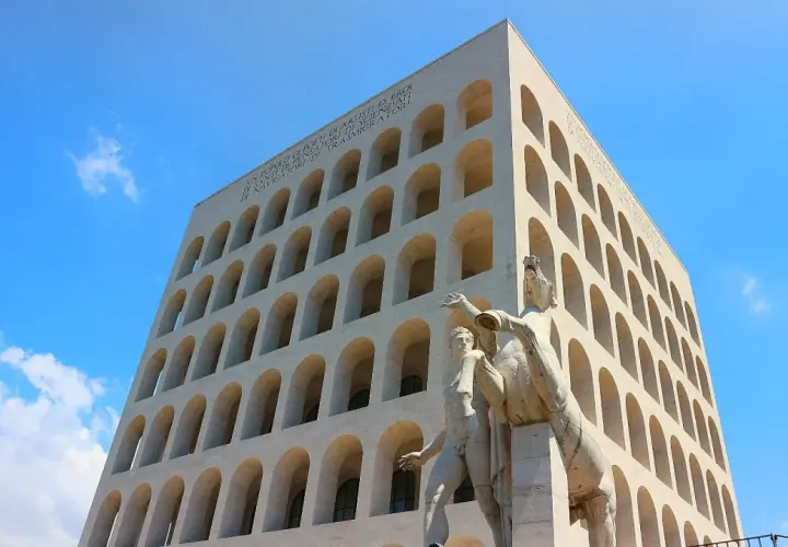 Palazzo Mussolini, con la sua facciata ad archi sovrapposti, domina il cielo azzurro. Una scultura di uomini e cavalli si erge in primo piano.