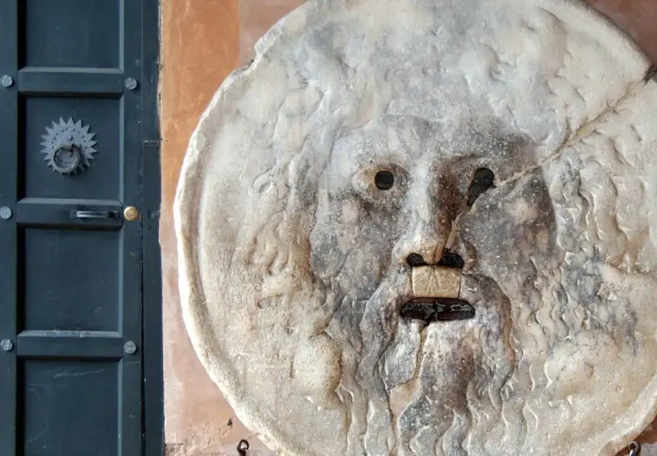 La Bocca della Verità, un volto scolpito nella pietra, sembra pronto a giudicare chiunque osi mentire. Accanto, una porta scura.