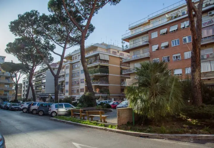 Edifici residenziali e pini marittimi nel quartiere Balduina a Roma. Auto parcheggiate lungo la strada, panchine e vegetazione rigogliosa.