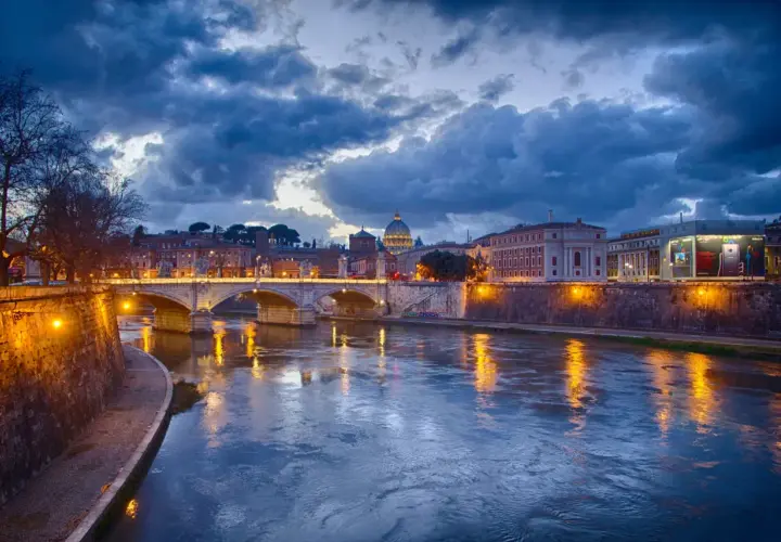 Il Tevere riflette le luci di Roma al tramonto, con la cupola di San Pietro sullo sfondo. La **Trastevere nightlife** è già nell'aria, promessa di serate vivaci.