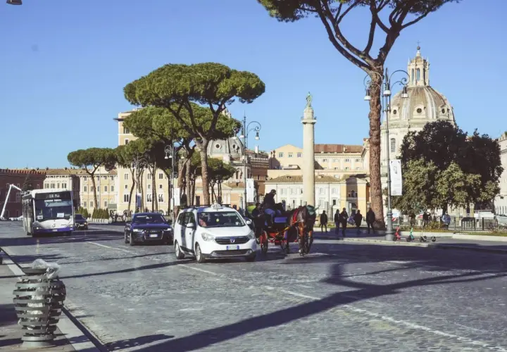 Un bus metro, taxi e carrozza a cavalli sfrecciano su un acciottolato a Roma, con edifici storici e pini marittimi sullo sfondo.