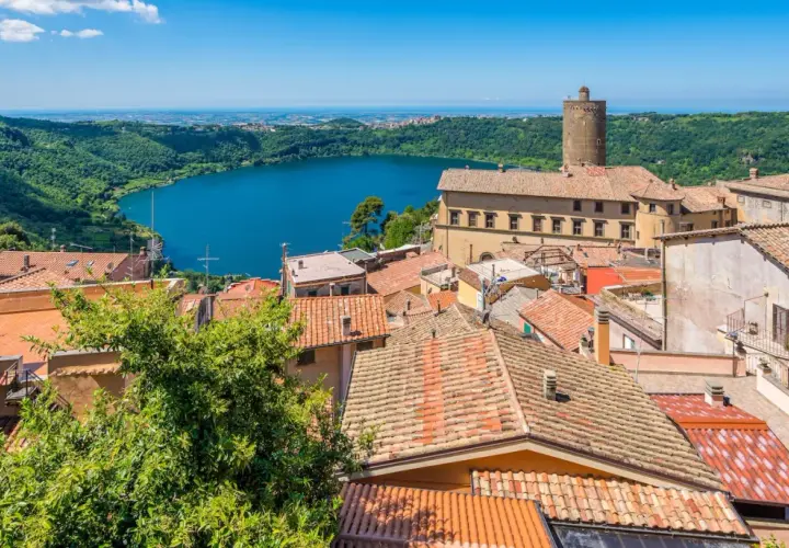 Panoramica dei colli di Roma oggi: un lago azzurro circondato da colline verdi e un borgo con tetti rossi e una torre medievale.