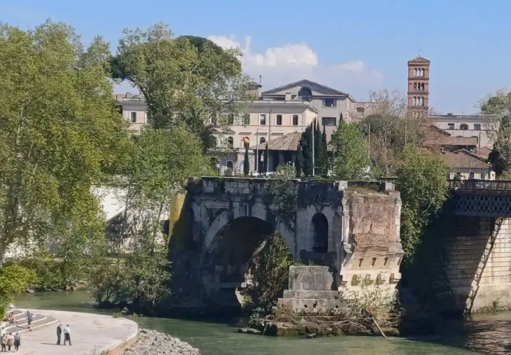 Antico **ponte rotto a Roma**, con rovine e vegetazione che si arrampicano sui resti. In lontananza, edifici storici e una torre campanaria.