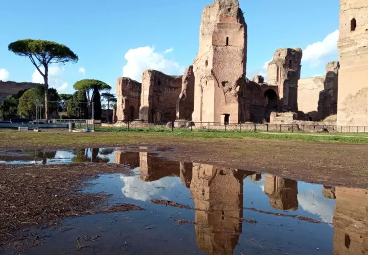 Le maestose rovine delle **Terme di Caracalla** si specchiano in una pozza d'acqua, sotto un cielo azzurro con nuvole bianche.