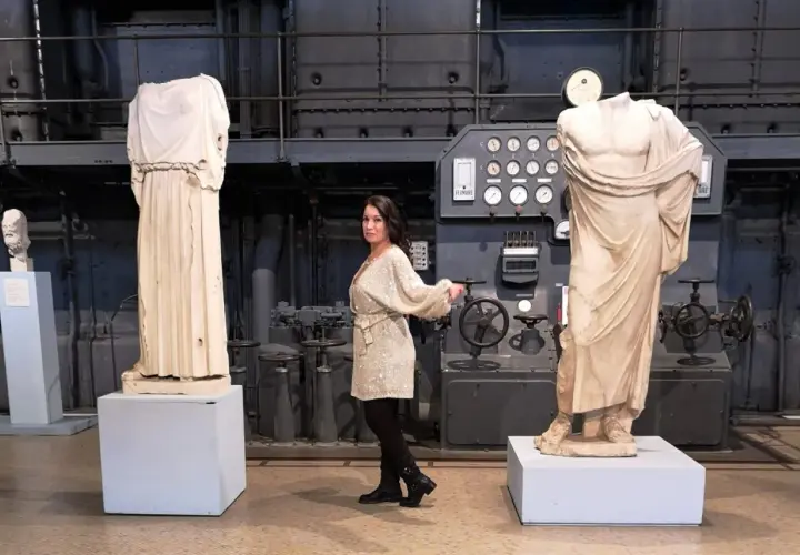 Donna in abito scintillante tra statue classiche e macchinari industriali, forse nel vecchio gasometro.