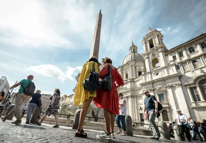 Turisti ammirano l'obelisco e la chiesa a Roma ad agosto, con il sole che illumina la piazza.