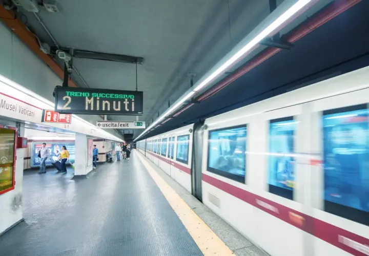 Stazione metro Roma: un treno in arrivo tra 2 minuti. Persone attendono sul binario, un cartello indica "uscita/exit".