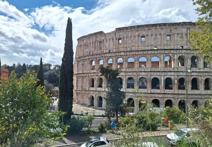Le maestose **colonne di Roma** del Colosseo si stagliano contro un cielo azzurro con nuvole bianche. Alberi verdi e un'auto bianca completano la scena.