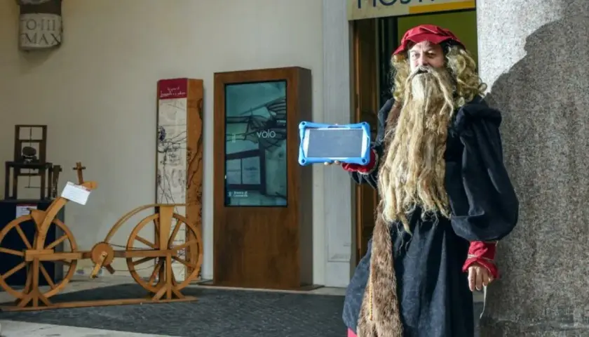 Uomo vestito da Leonardo da Vinci con tablet, pronto per una caccia al tesoro a Roma, vicino a un'antica bicicletta di legno.
